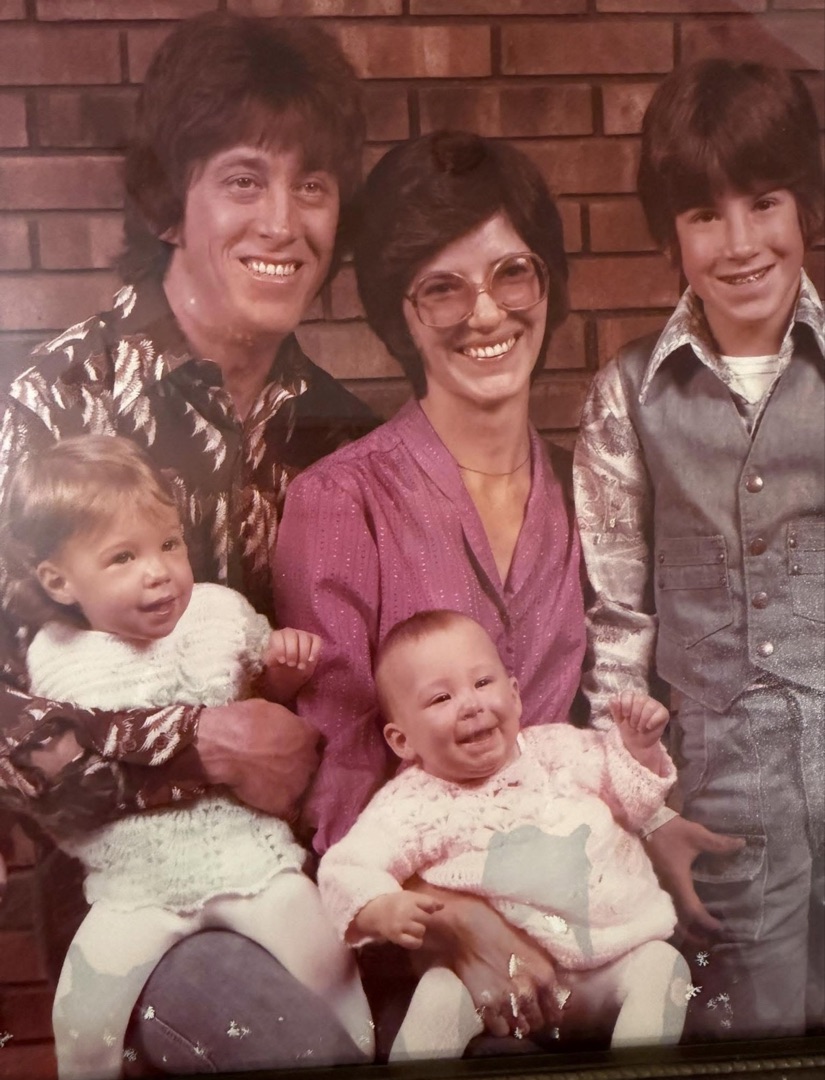 A family portrait from the 1970s — young parents smiling with their three kids in front of a brick wall.